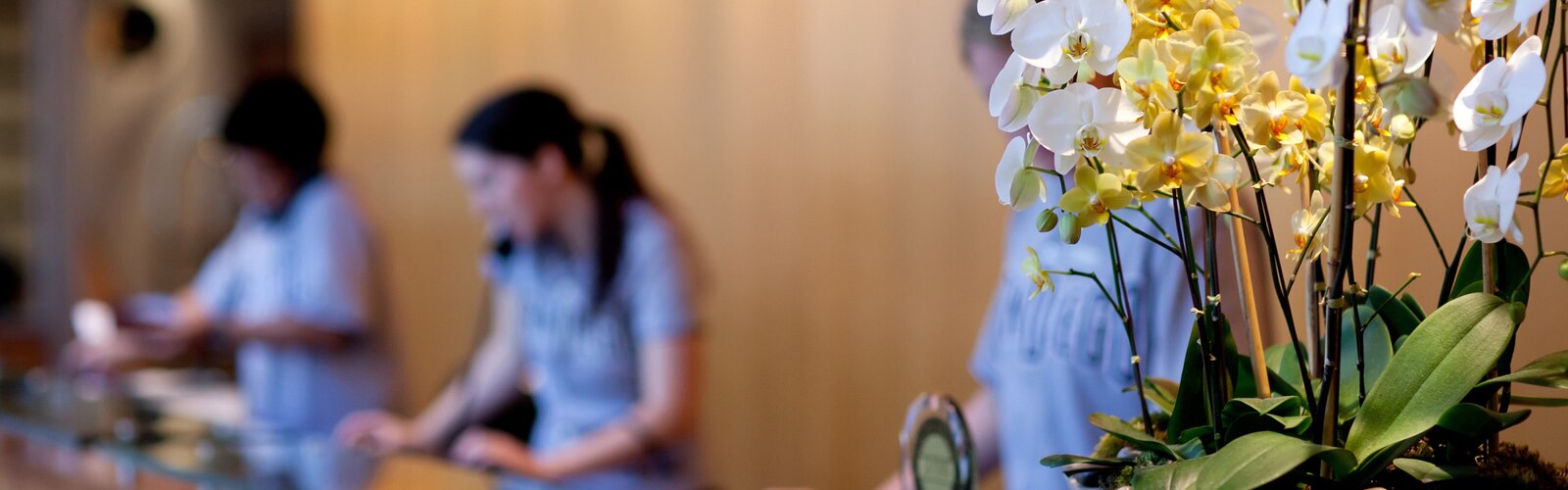 Un pot d’orchidée à la réception d’un hôtel, avec trois réceptionnistes en arrière-plan