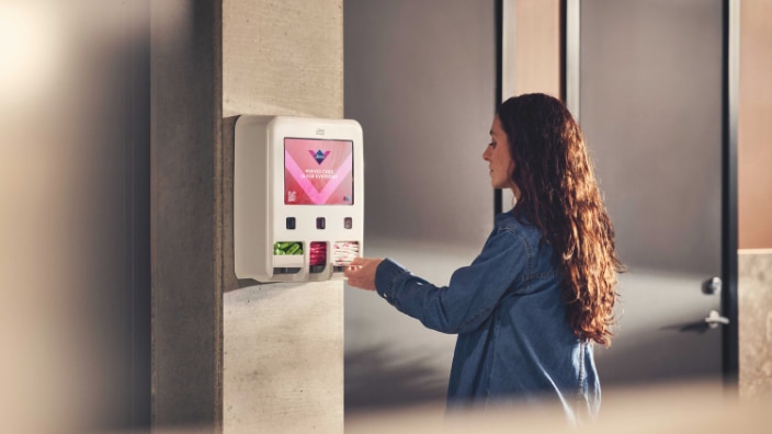 Person som använder en väggmonterad dispenser för mensskydd och väljer en mensprodukt från dispensern. Person som använder en väggmonterad dispenser för mensskydd och väljer en mensprodukt från dispensern.