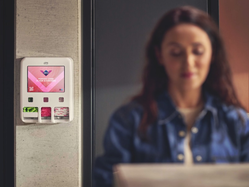 Väggmonterad dispenser för mensskydd med mensprodukter, med en person suddigt i förgrunden. Väggmonterad dispenser för mensskydd med mensprodukter, med en person suddigt i förgrunden.